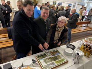 Teamarbeit war auch das Anschneiden der Fototorte, die mit Fotos des Waldbahnstreckennetzes versehen war. Landrat Sebastian Gruber (v.li.), Staatsminister Christian Bernreiter und Landrätin Rita Röhrl schnitten den Kuchen an. Foto: Heiko Langer/Landkreis Regen